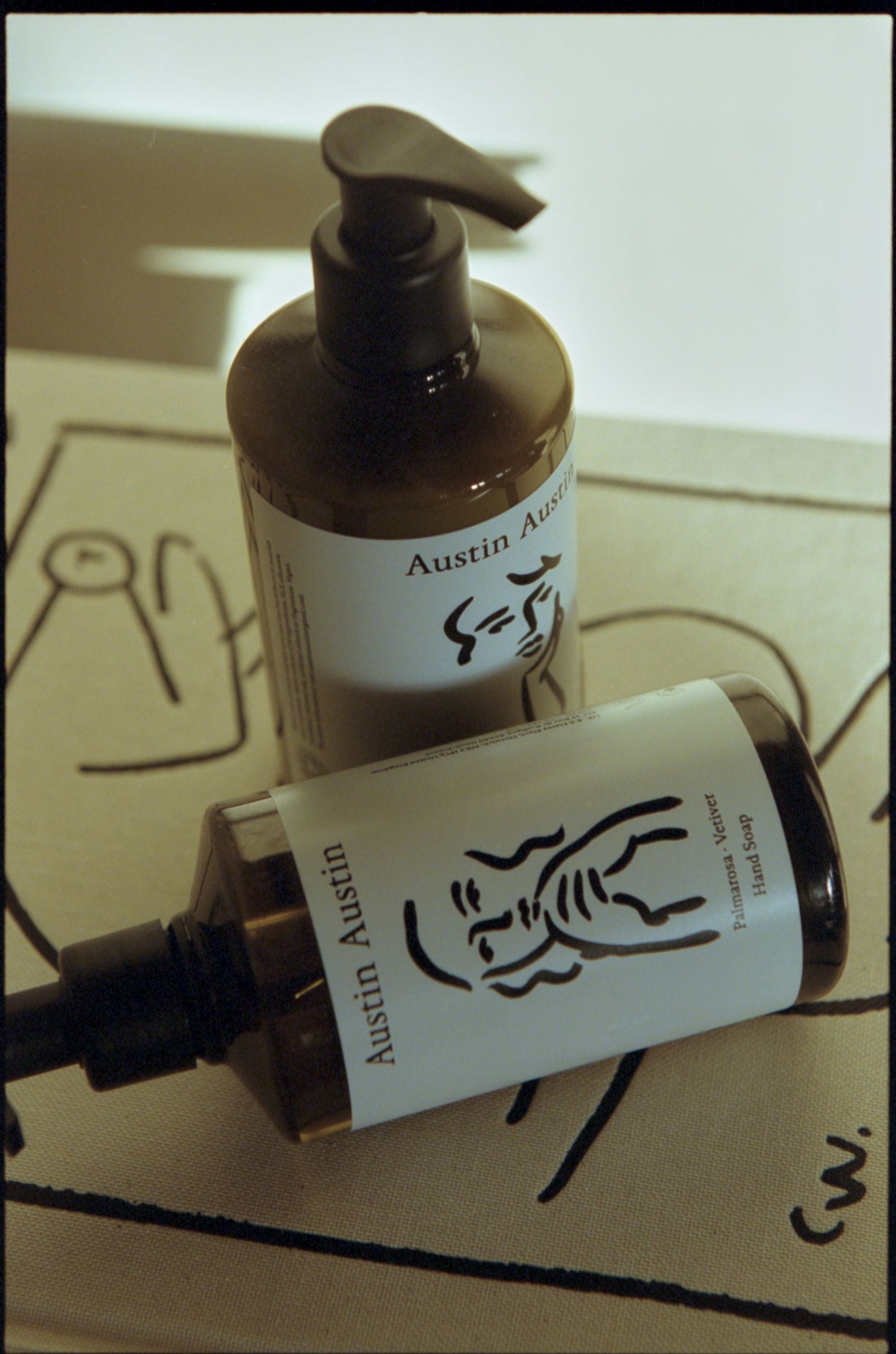 Austin Austin Palmarosa and Vetiver Hand Soap 300ml amber bottle with black pump dispenser placed alongside another bottle, Christian Newby figurative artwork visible on both label and box