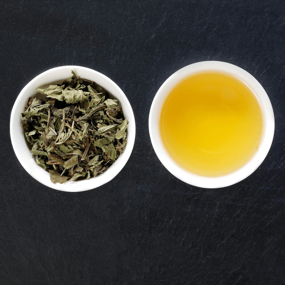 Good & Proper Tea peppermint herbal tea with dried whole leaves in a bowl and a cup of brewed golden tea on a dark slate surface