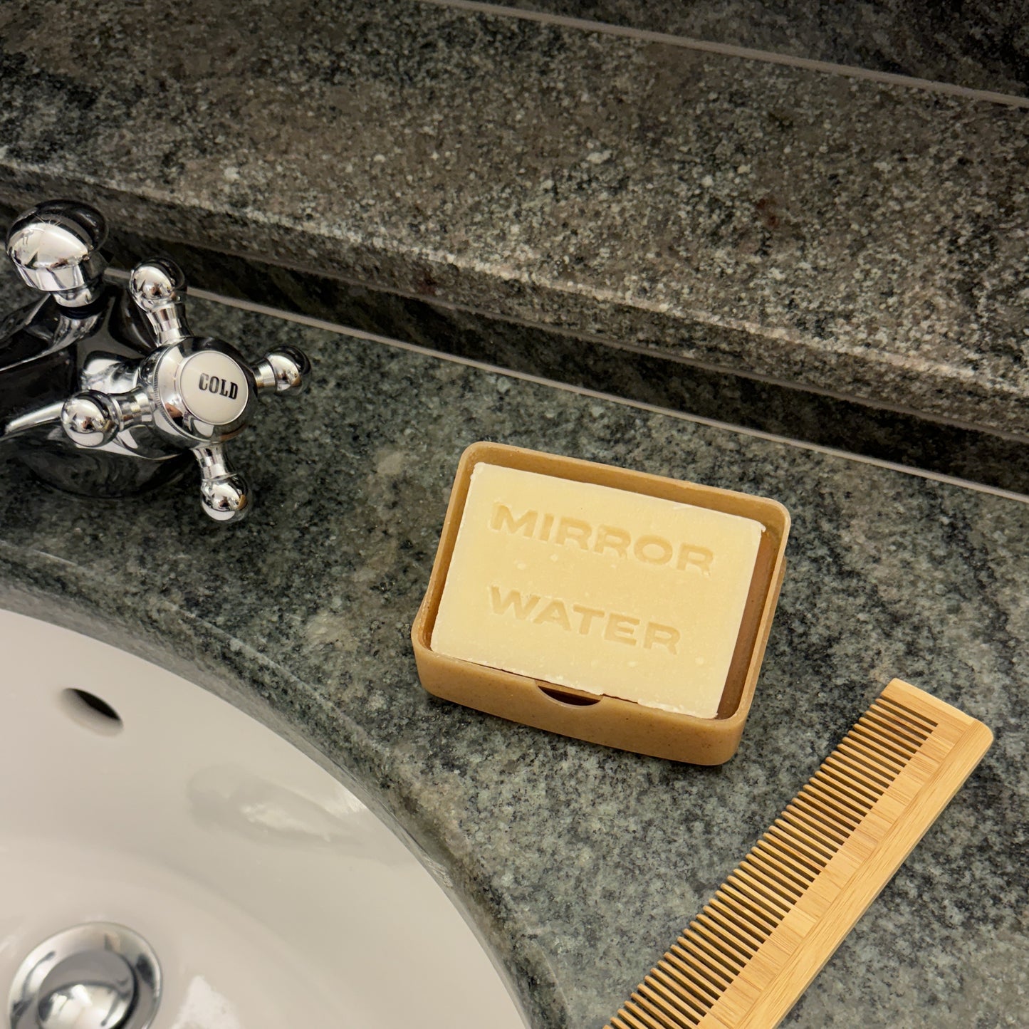 Mirror Water Shea Butter Brick solid face and body cleanser bar in a tray next to a wooden comb on a granite bathroom vanity