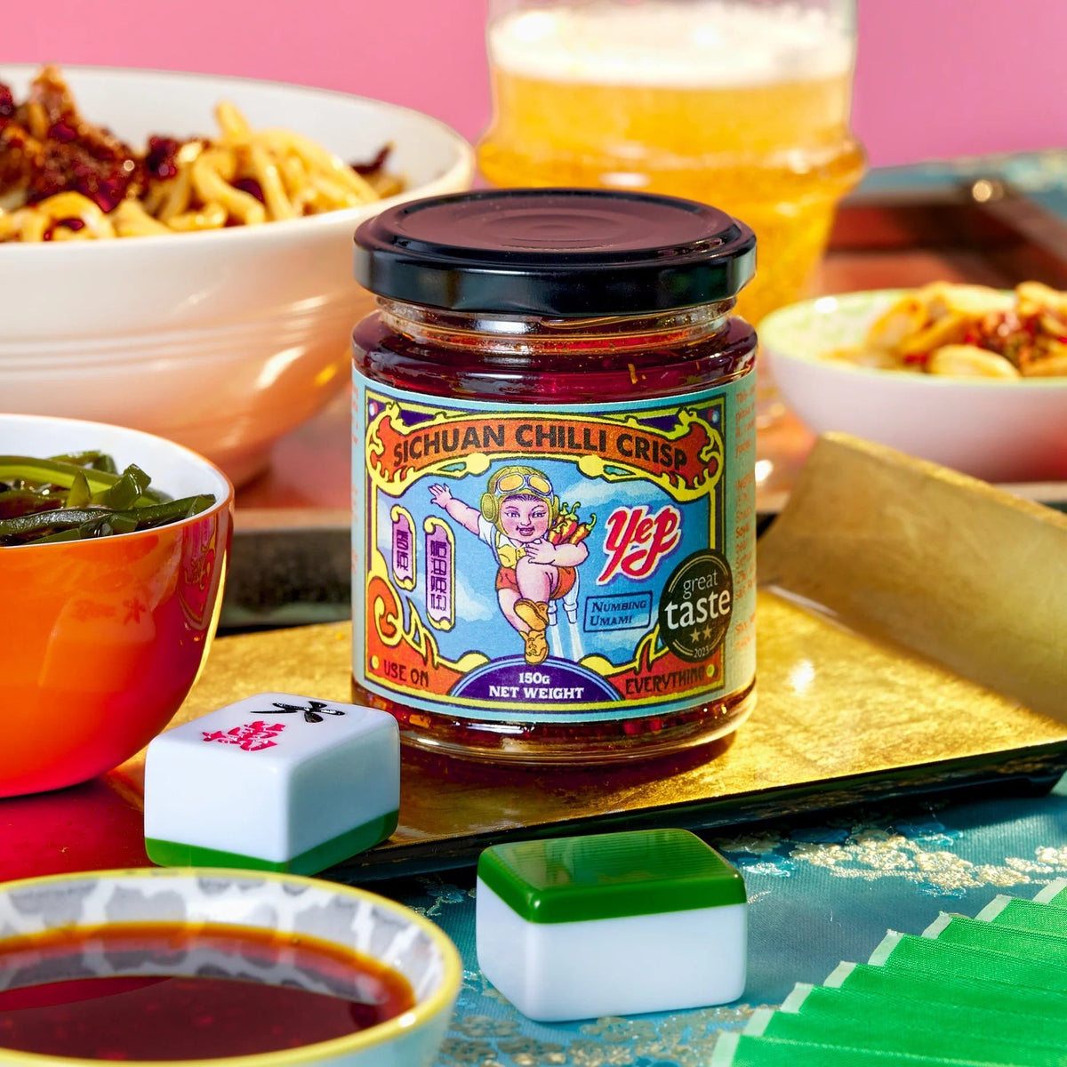 Yep Kitchen Sichuan Chilli Crisp oil jar with vibrant illustrated label, surrounded by bowls of noodles and mahjong tiles.
