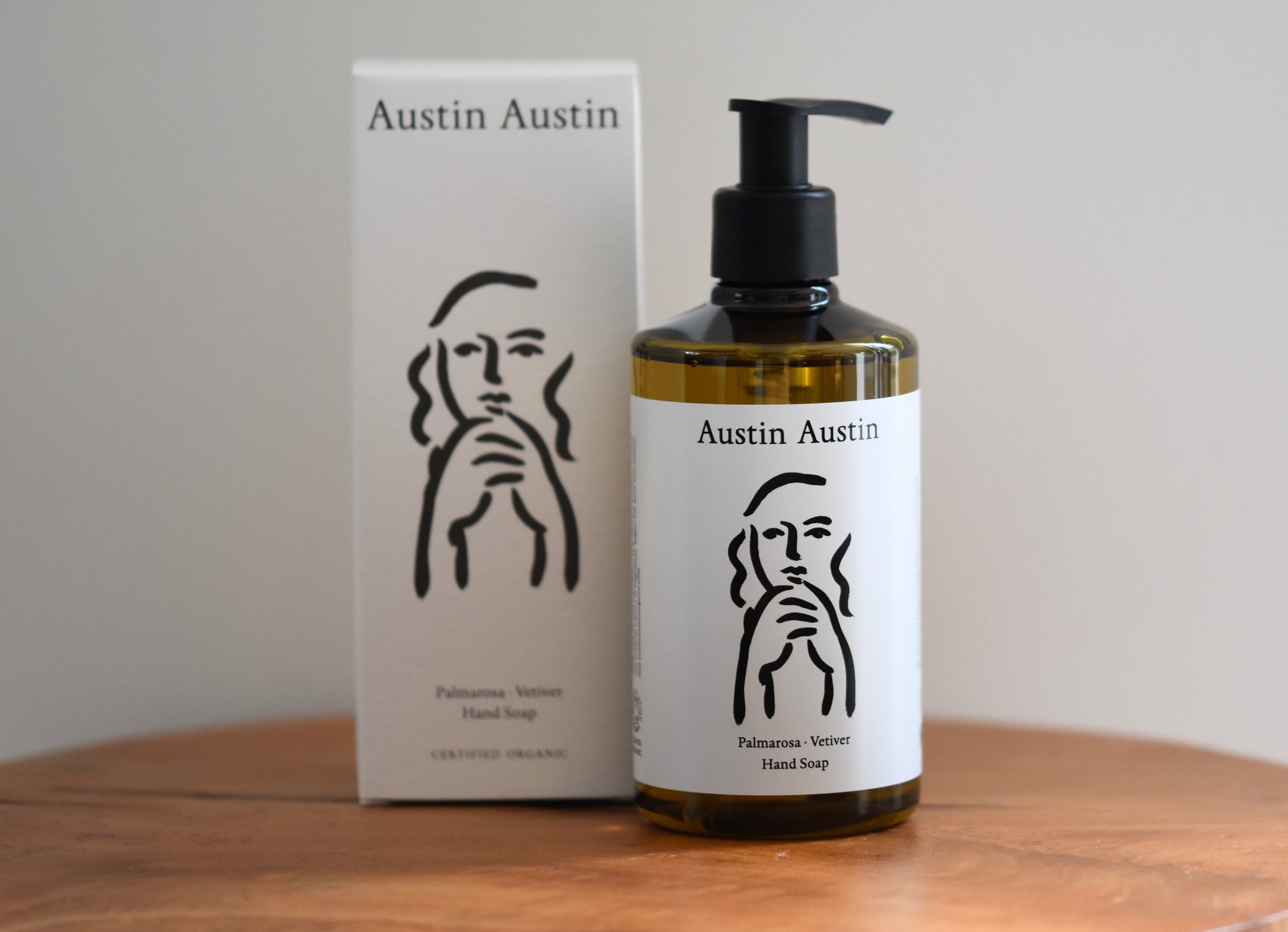 Austin Austin Palmarosa and Vetiver Hand Soap 300ml bottle and white box displayed on a round wooden side table, warm natural lighting showing the amber tone of the soap through the bottle and the Indian ink label artwork