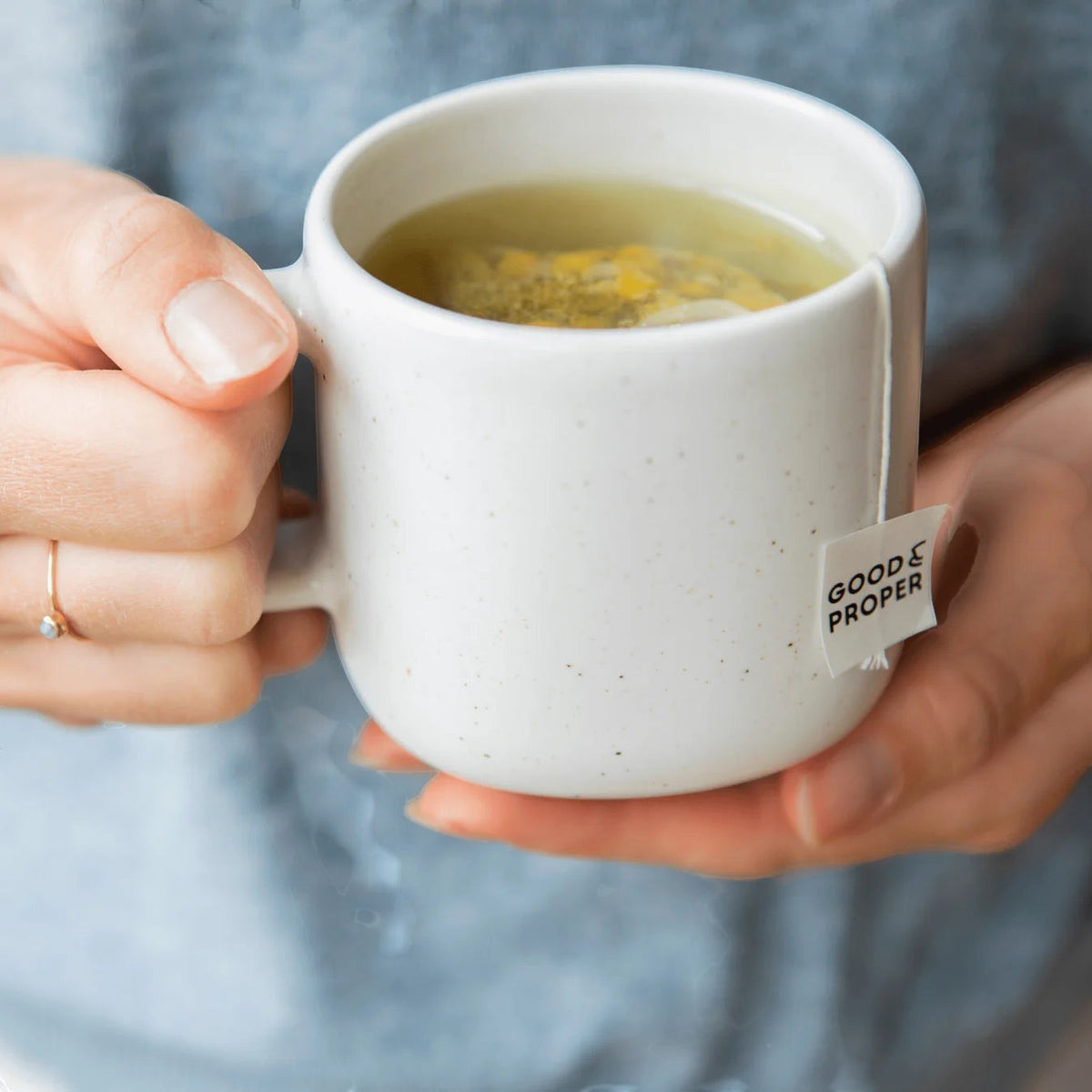 Good and Proper Tea  chamomile tea bag steeping in a white ceramic mug held by hands, showing the compostable tag and floral tea.