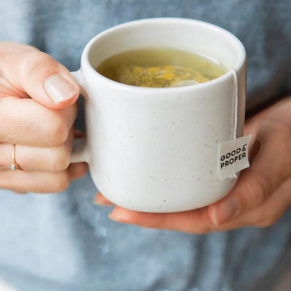 Good and Proper Tea  chamomile tea bag steeping in a white ceramic mug held by hands, showing the compostable tag and floral tea.