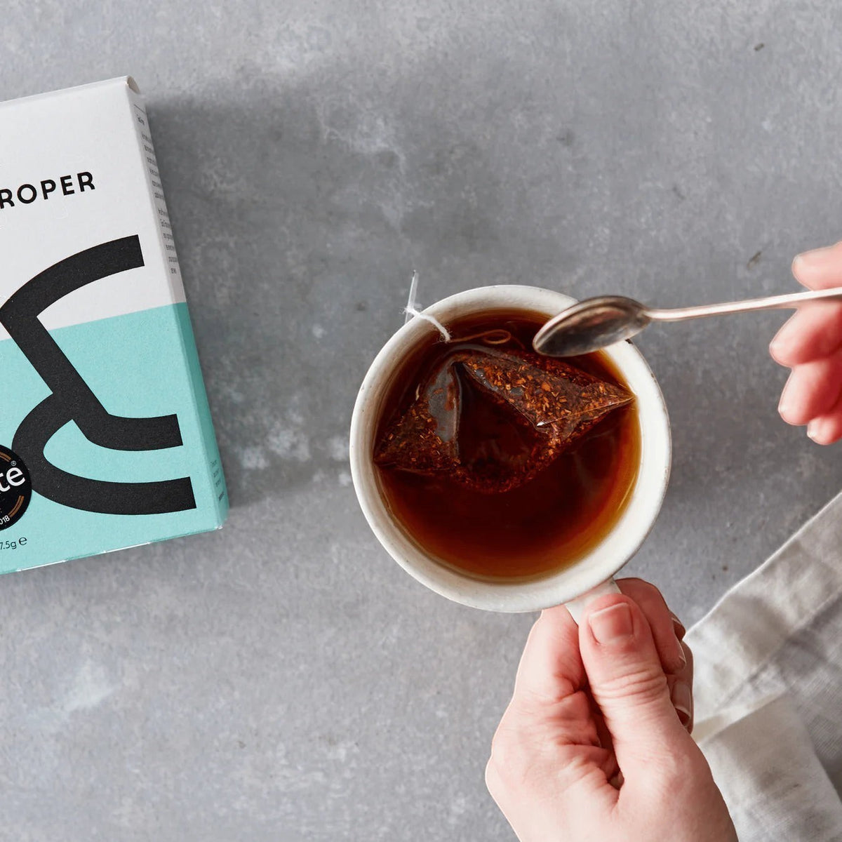 Good and Proper Tea rooibos tea bag steeping in a white cup with a spoon, next to a recyclable cardboard box on a grey surface.