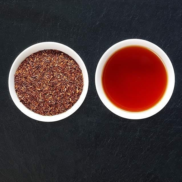 Good and Proper Tea rooibos tea featuring a bowl of loose red tea leaves next to a cup of brewed amber tea on a dark slate surface