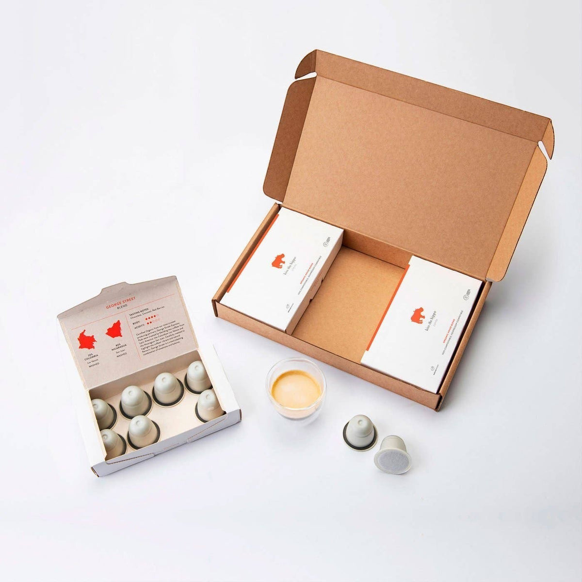 Kiss the Hippo organic coffee pods in compostable packaging with a small glass of espresso on a white background