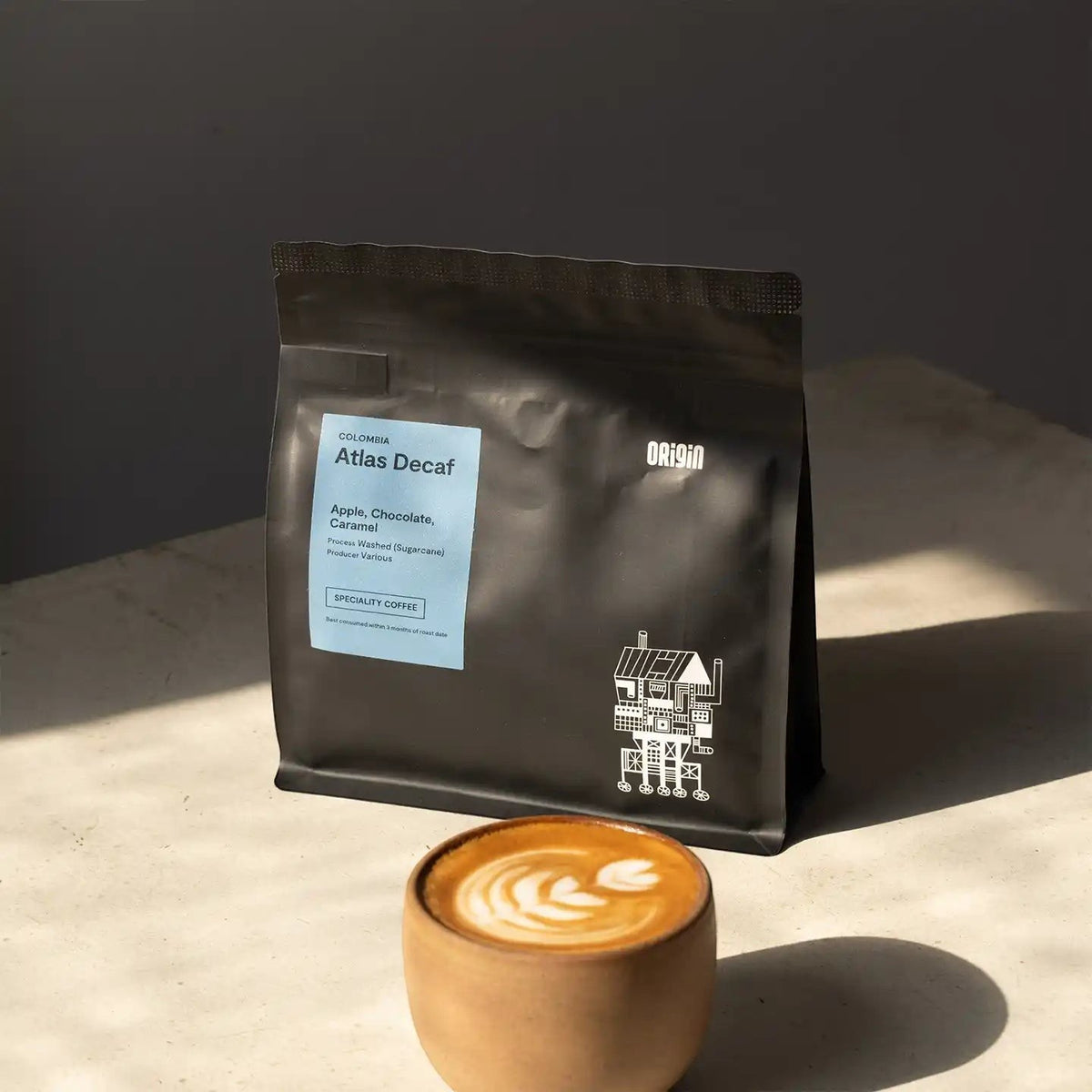 Origin Coffee Atlas Decaf coffee bag with a light blue label next to a ceramic cup of latte art on a sunlit surface.