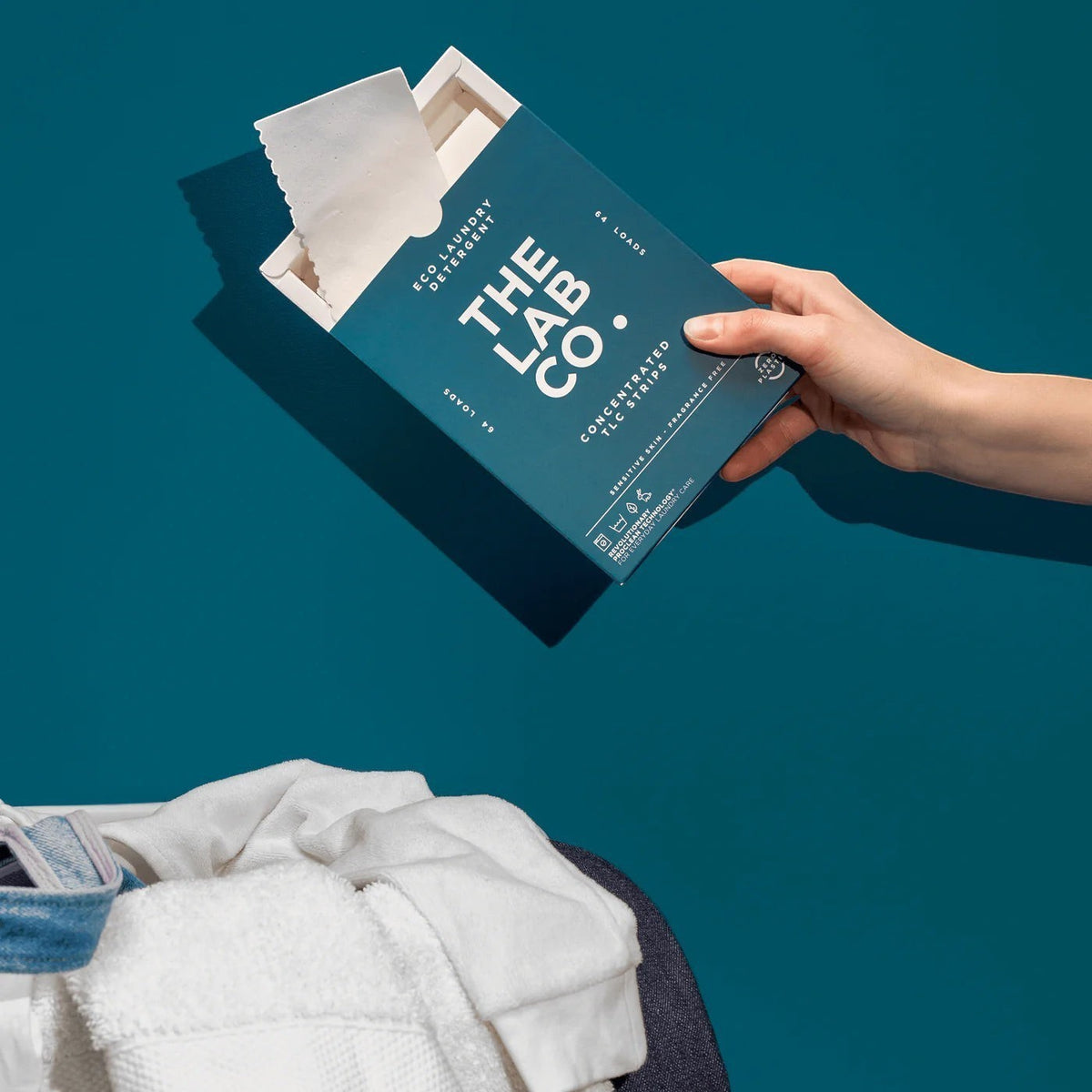 hand holding a box of The Lab Co. fragrance-free laundry detergent strips over a pile of white laundry