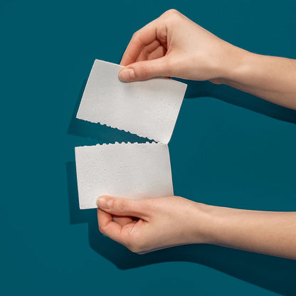 The Lab Co. fragrance-free laundry detergent strip being torn in half by hands against a dark teal background