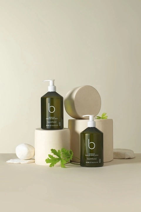 Bamford Geranium hand and body lotion in a dark green pump bottle displayed on minimalist beige pedestals.