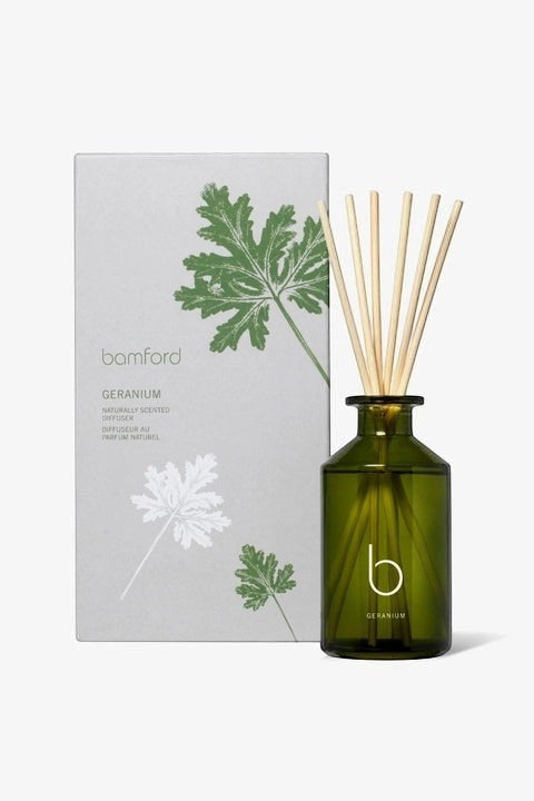 bamford Geranium diffuser in a green glass bottle with wooden reeds next to its botanical-print packaging box.