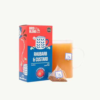 Bird and Blend Rhubarb & Custard rooibos tea box with a pyramid tea bag and a glass mug of brewed tea on a light background
