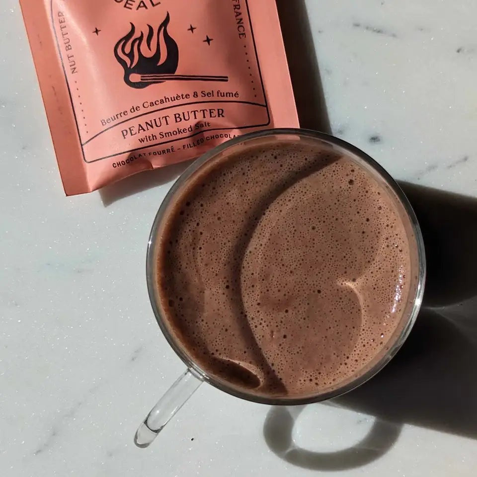 Cosmic Dealer Chai Cacao drinking chocolate in a glass mug next to a recyclable sachet on a marble surface