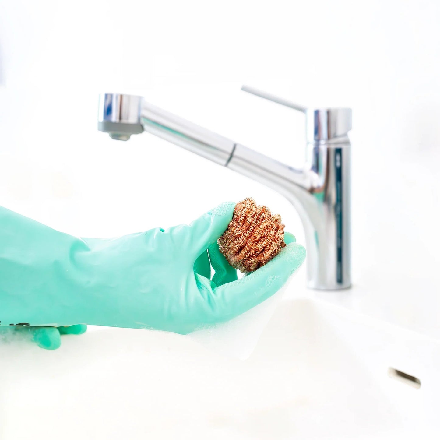 Seep eco-friendly copper scourer held by a hand in a mint green rubber glove near a kitchen sink faucet