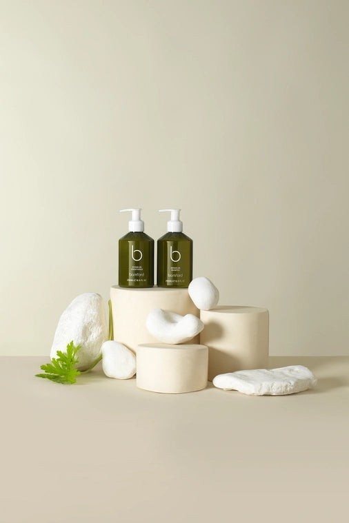 Bamford Geranium Shampoo bottles in dark green with pump dispensers, displayed on minimalist pedestals with white stones.