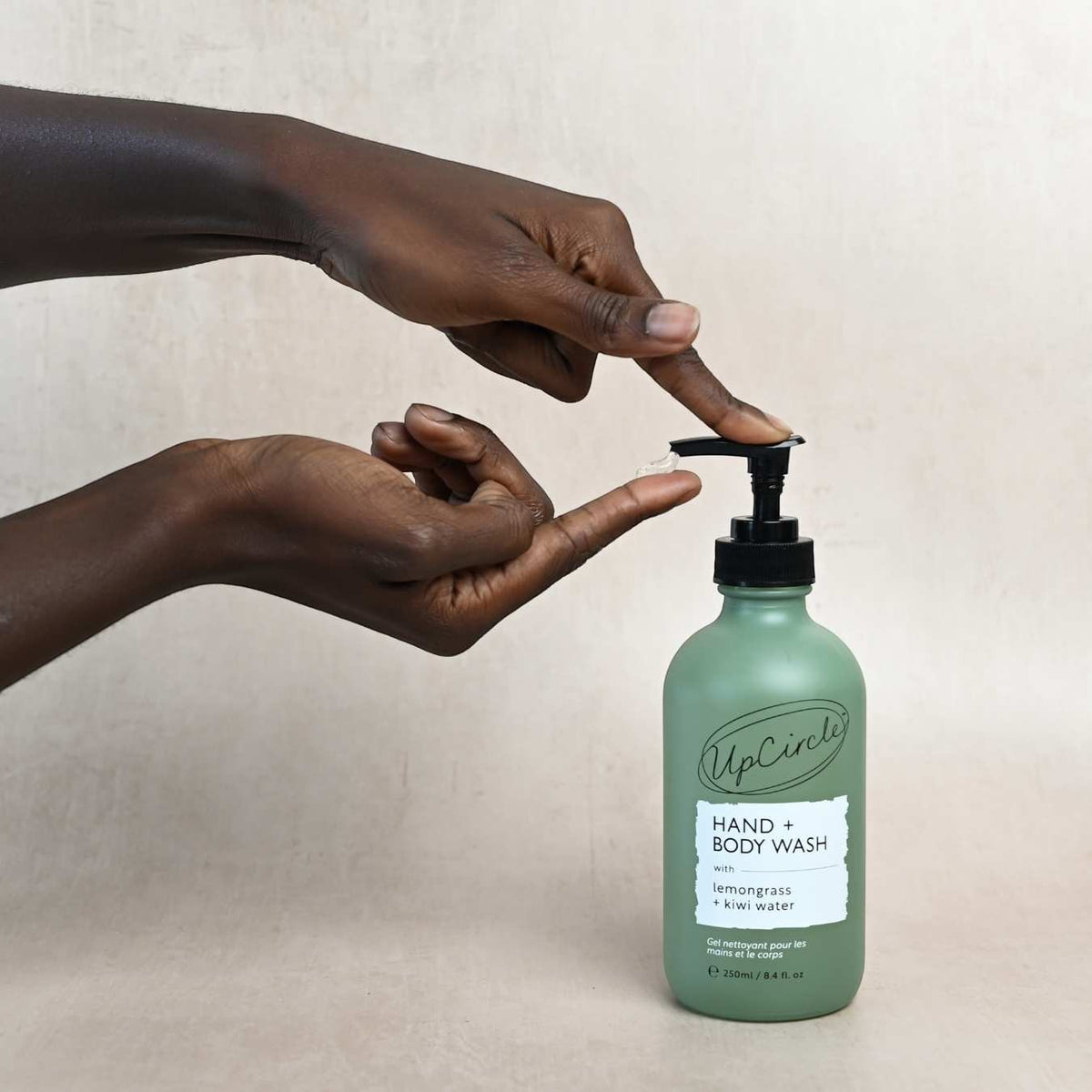 UpCircle hand and body wash in a frosted green glass bottle with pump, being dispensed by a person's hands.