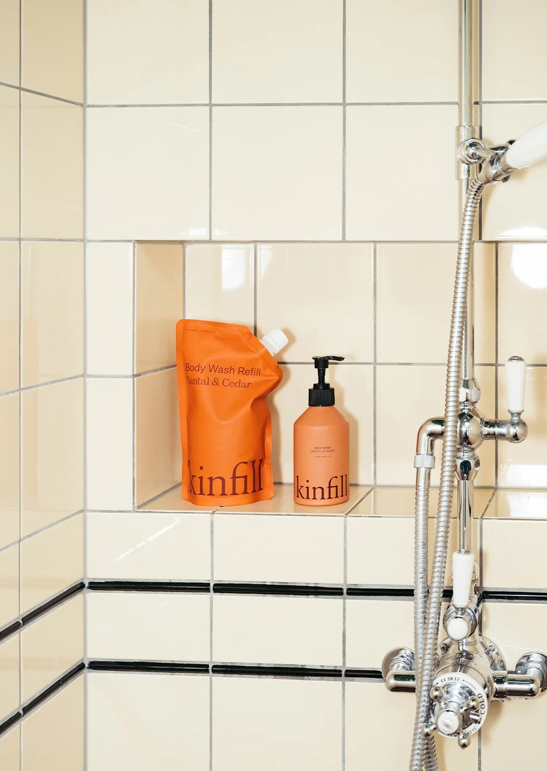 Kinfill body wash refill pouch and refillable aluminium pump bottle in a tiled shower niche.