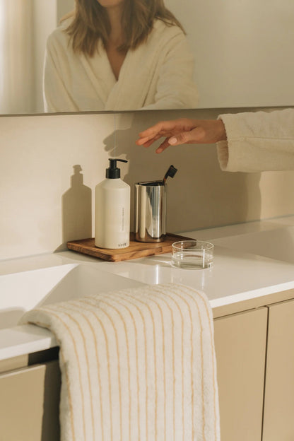Kinfill hand wash bottle on a wooden tray in a minimalist bathroom with a person reaching for the dispenser.