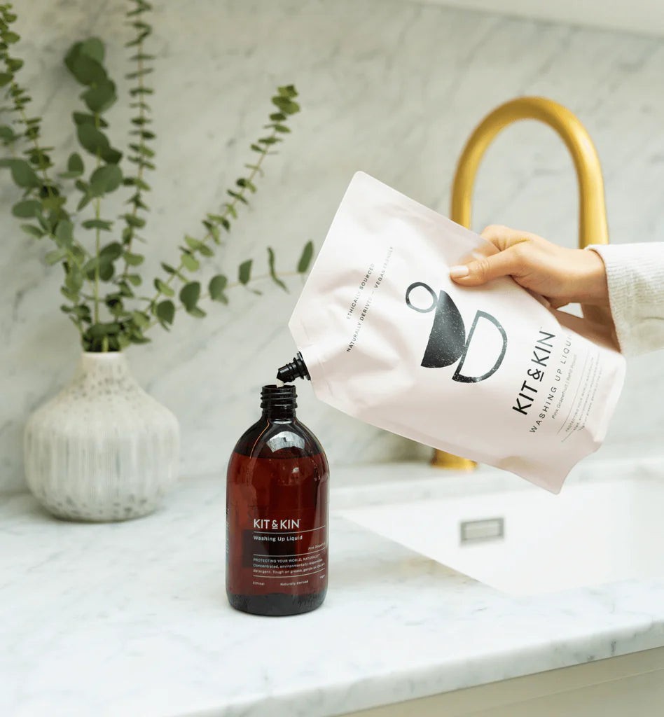 Kit & Kin eco-friendly washing-up liquid being poured from a refill pouch into a glass bottle in a kitchen.