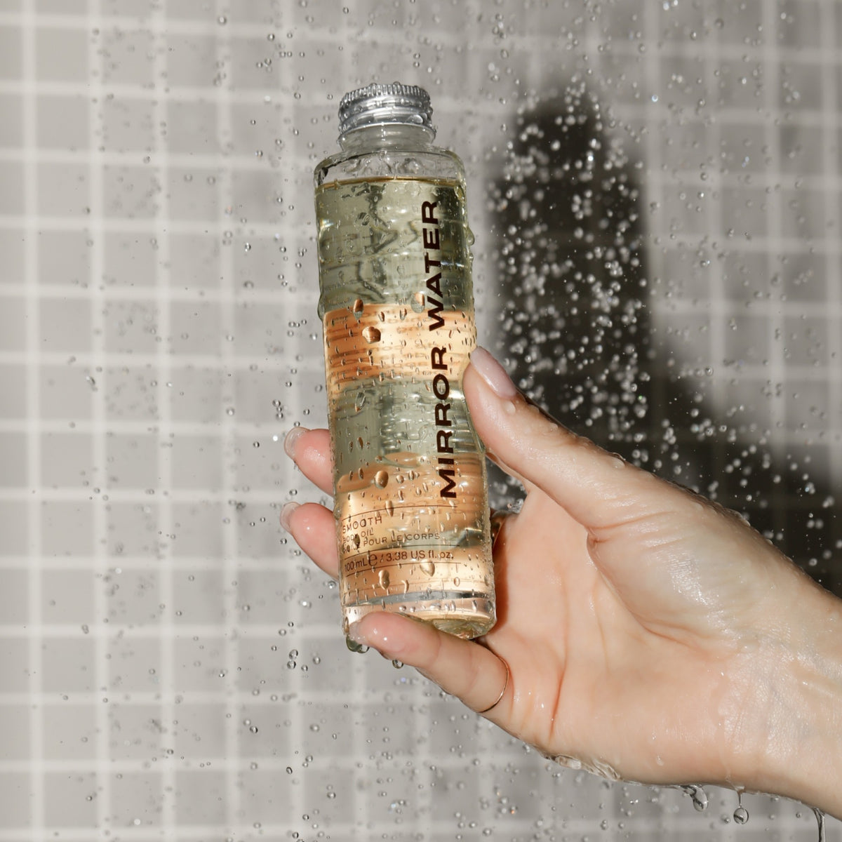 Mirror Water body oil in a clear 100ml glass bottle held under a shower spray against a tiled wall