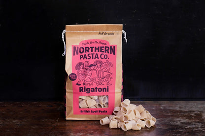 Northern Pasta Co. Rigatoni in a brown paper bag with a pile of bronze-extruded spelt pasta on a wooden surface.