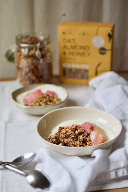 Nibs Etc. oat, almond, and honey granola served in bowls with yogurt and poached rhubarb on a white cloth.
