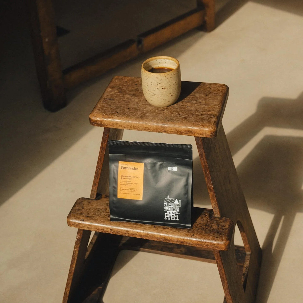 Origin Coffee Pathfinder whole bean coffee bag and a ceramic cup on a rustic wooden step stool in natural sunlight.