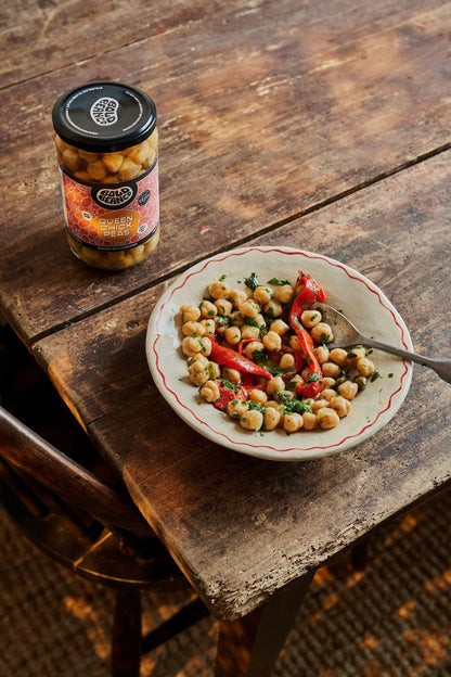 Bold Bean Co Queen Chickpeas jar next to a rustic bowl of prepared heirloom chickpeas with roasted red peppers and herbs.