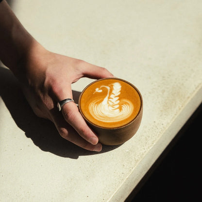 Origin Coffee Resolute coffee in a ceramic cup with swan latte art held by a hand on a sunlit stone surface