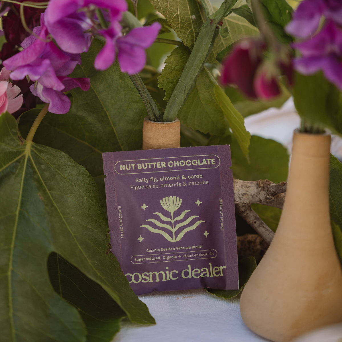 Cosmic Dealer Salty Fig Chocolate bar in purple packaging with almond, carob, and fig, displayed near purple flowers.