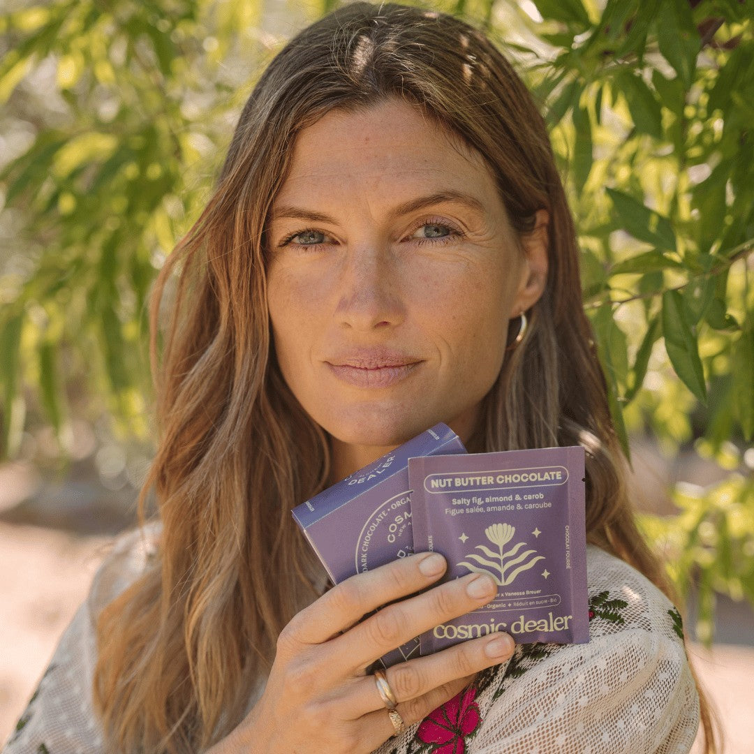 Cosmic Dealer model holding two purple Salty Fig Chocolate bars with almond butter and carob in a sunlit garden setting.