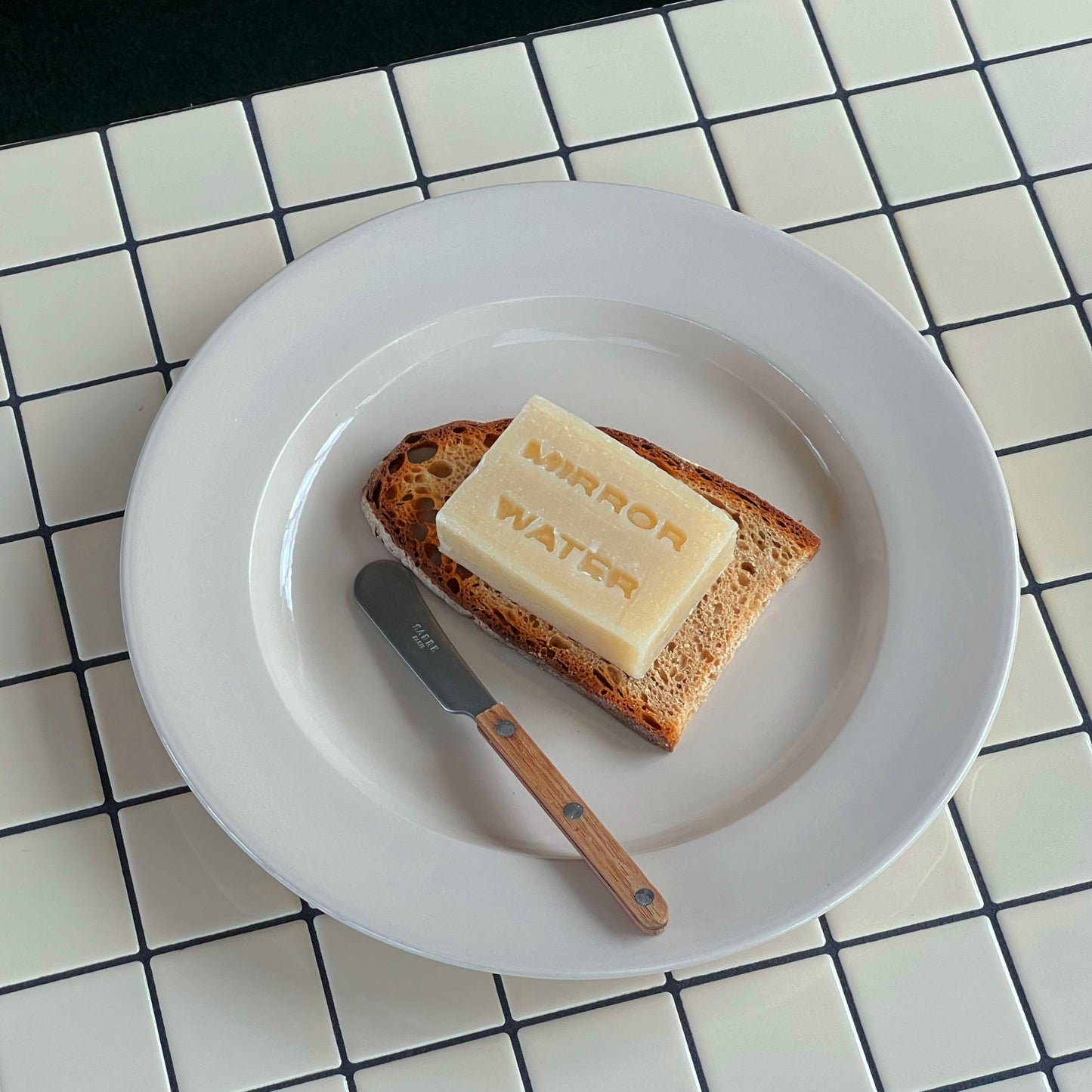 Mirror Water Shea Butter Brick solid cleanser bar resting on a slice of toasted bread with a small butter knife on a plate.