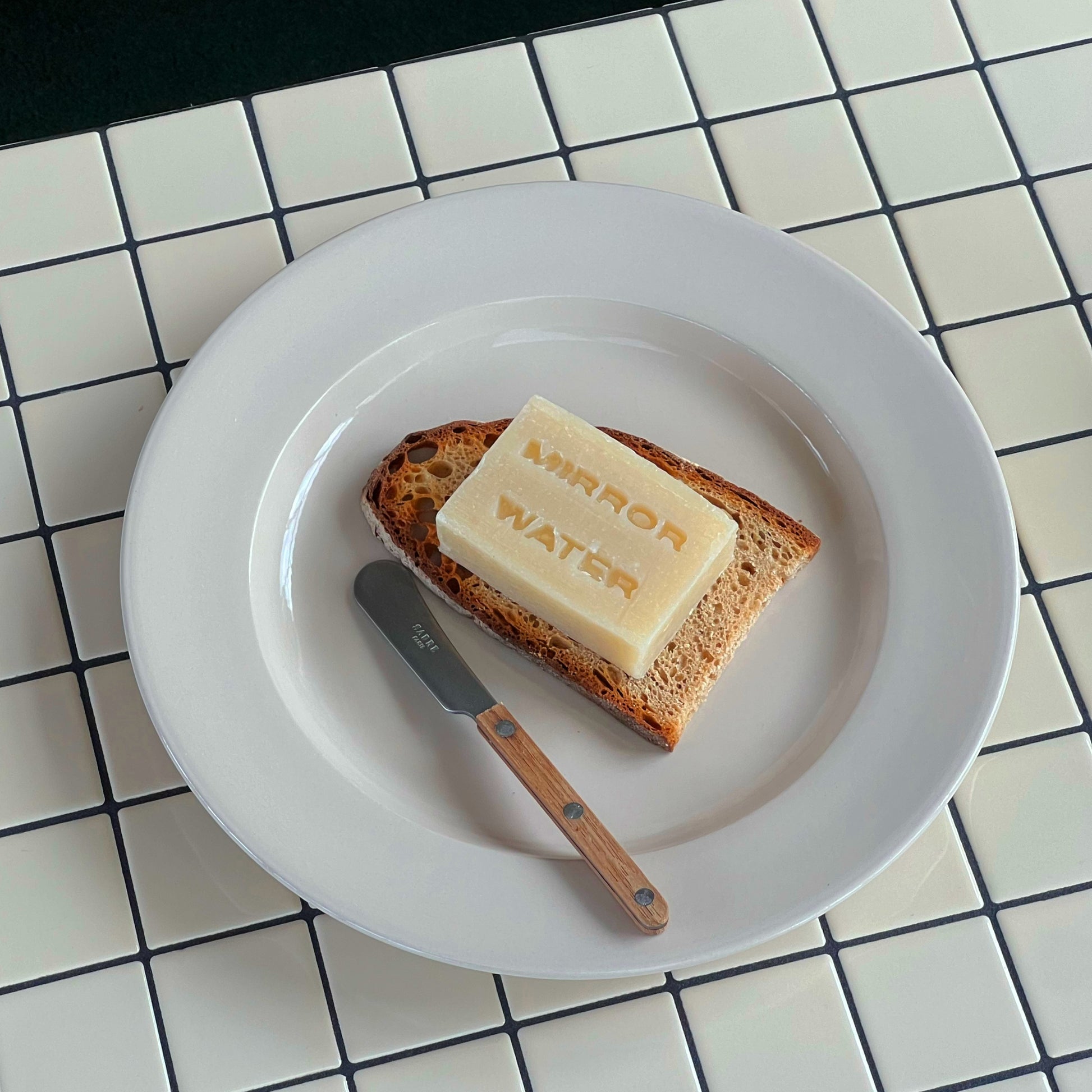 Mirror Water Shea Butter Brick solid cleanser bar resting on a slice of toasted bread with a small butter knife on a plate.