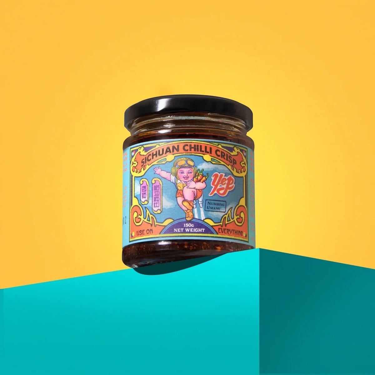 Yep Kitchen Sichuan Chilli Crisp oil in a glass jar with a colourful illustrated label on a teal block and yellow background