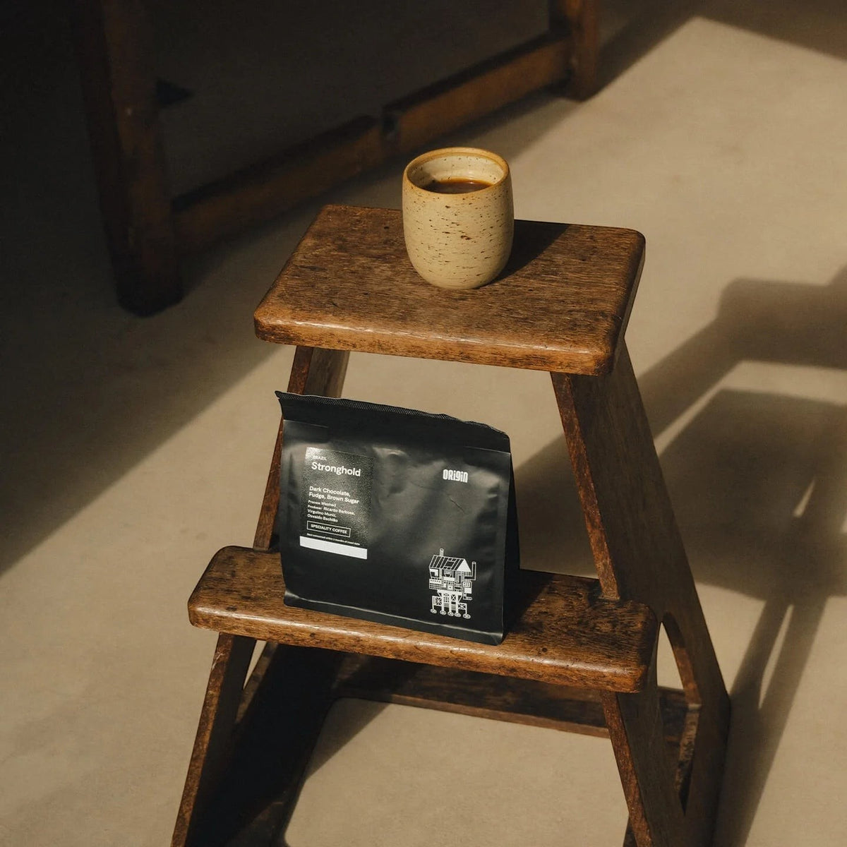 Origin Coffee Stronghold whole bean coffee bag and a ceramic cup on a rustic wooden step stool in soft natural light