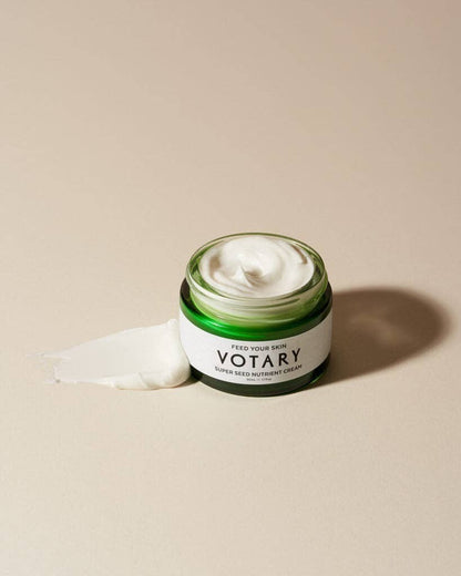 Votary Super Seed Nutrient Cream in a green glass jar with a dollop of white face cream on a neutral background.