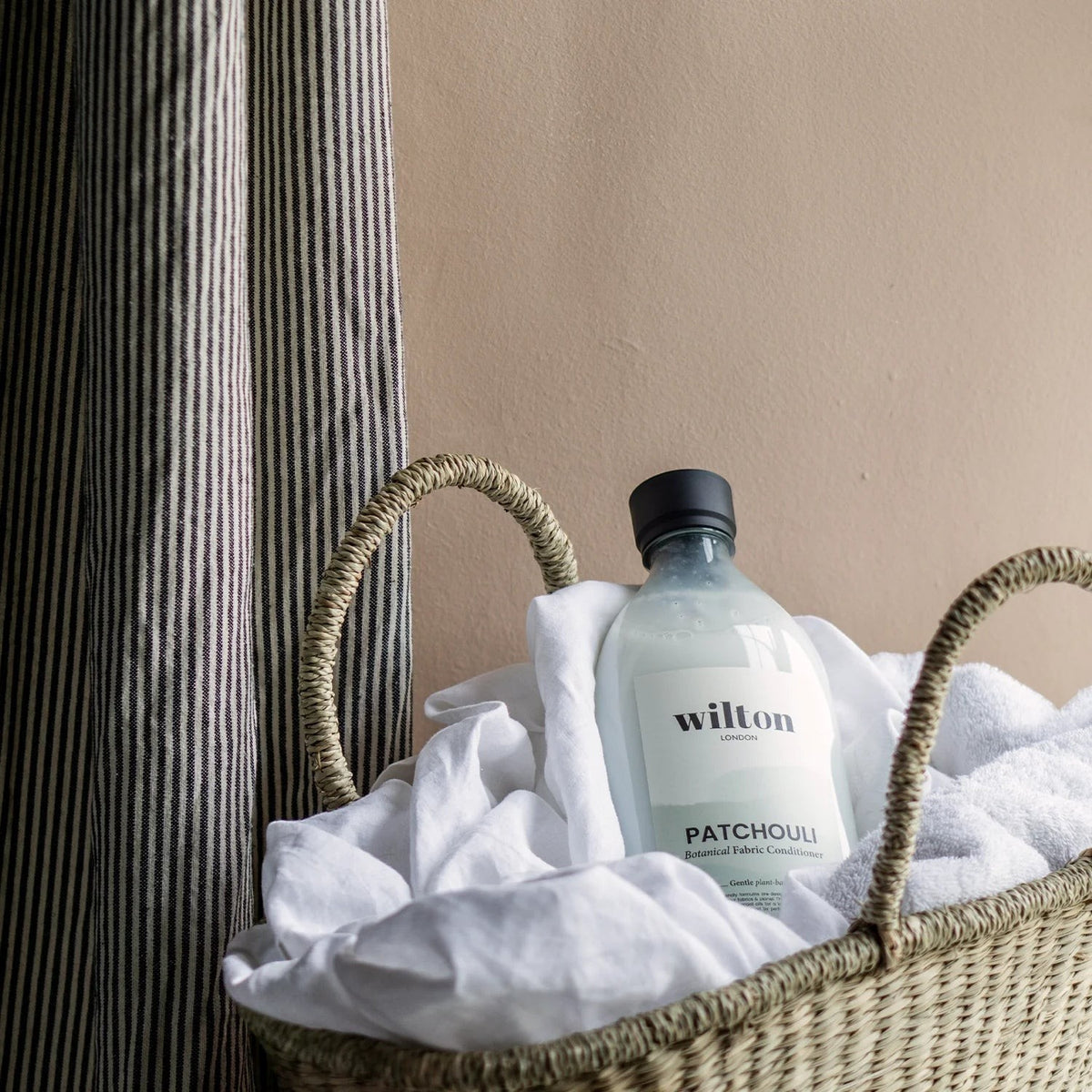 Wilton London Patchouli fabric conditioner bottle in a woven basket with white linens and striped curtains.