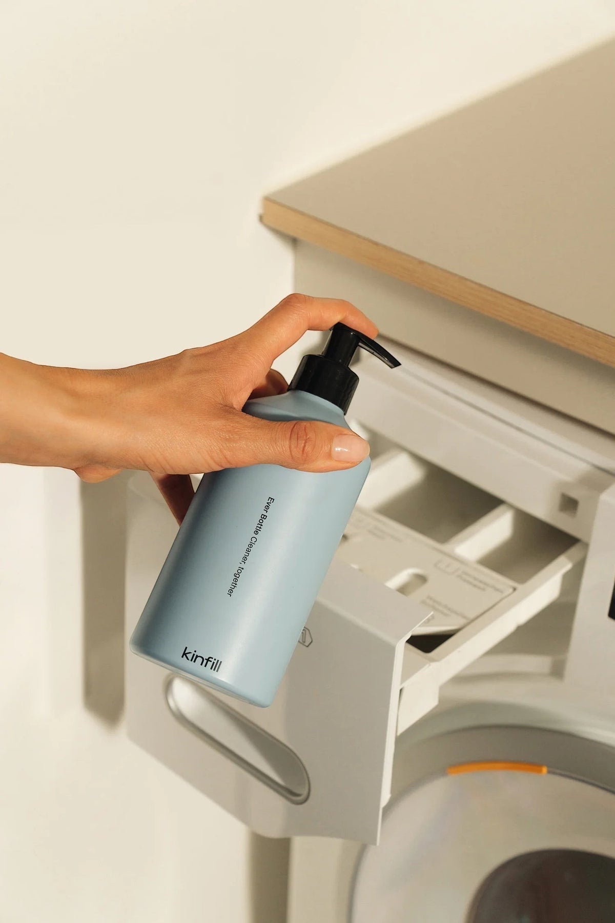 hand holding a light blue Kinfill refillable laundry detergent bottle next to a washing machine drawer
