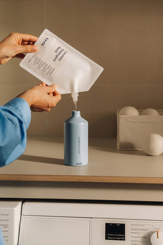 Kinfill laundry detergent refill pouch being poured into a light blue reusable bottle on a laundry room counter.