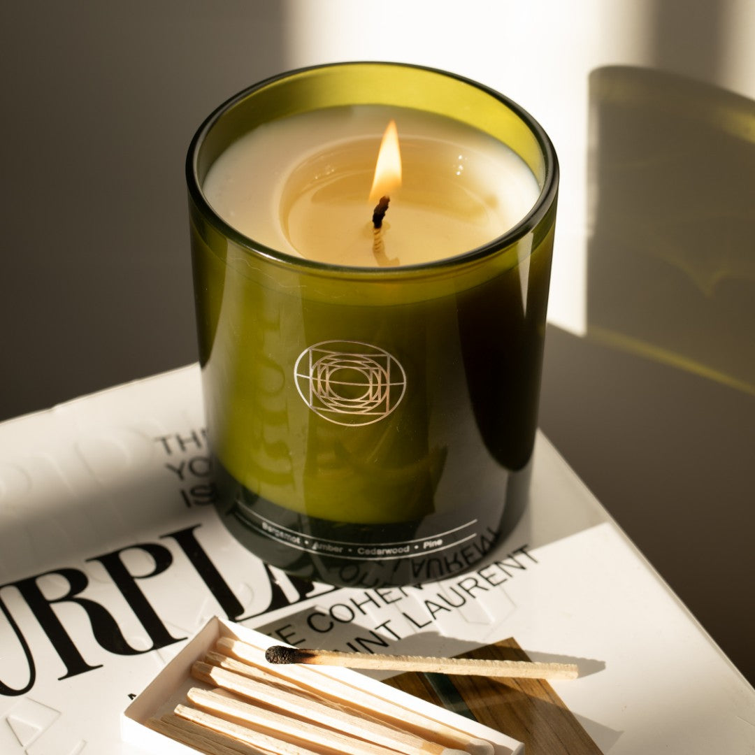 Mirror Water Sun Through Trees soy wax candle in a green glass jar with a lit wick, placed on a book with matches.