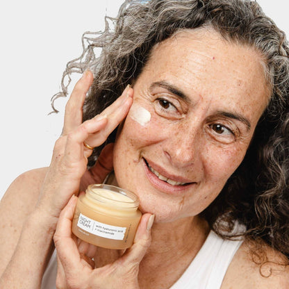 upcircle anti-ageing night cream in a glass jar held by a smiling woman applying the rich cream to her cheek