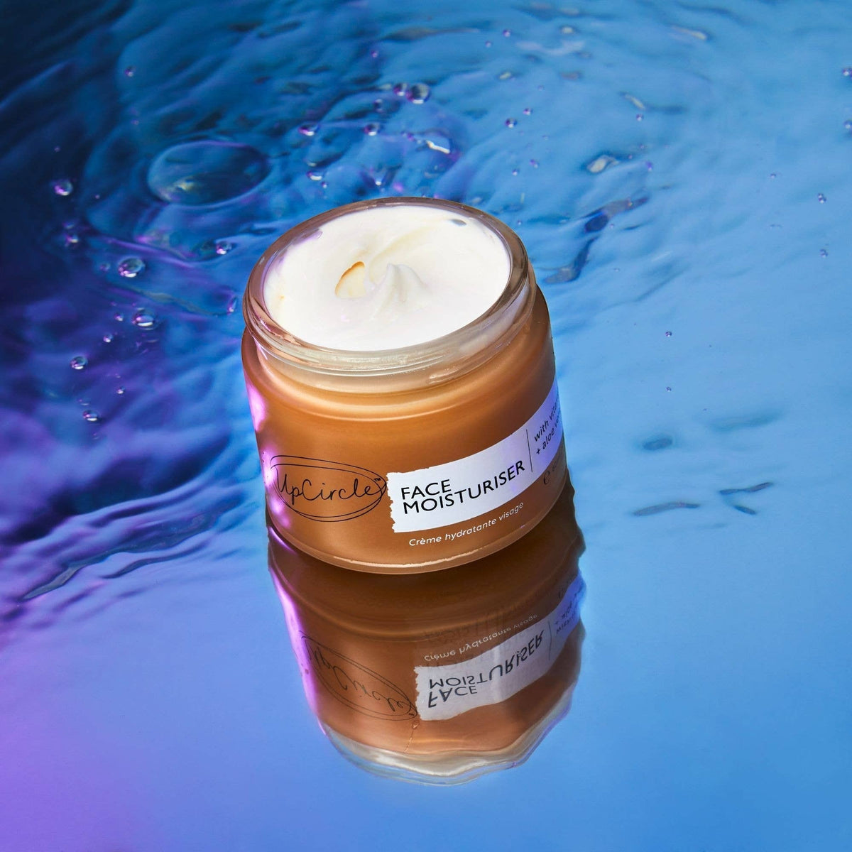 upcircle face moisturiser in a glass jar with a white cream texture, set against a reflective blue water background.