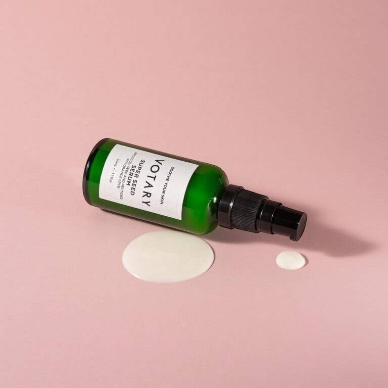 Votary Super Seed Serum in a green glass bottle with a pump, shown lying down with a drop of creamy serum on pink