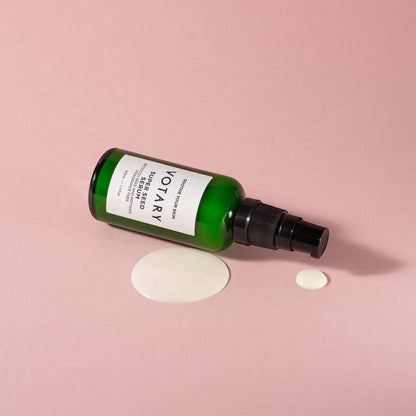 Votary Super Seed Serum in a green glass bottle with a pump, shown lying down with a drop of creamy serum on pink